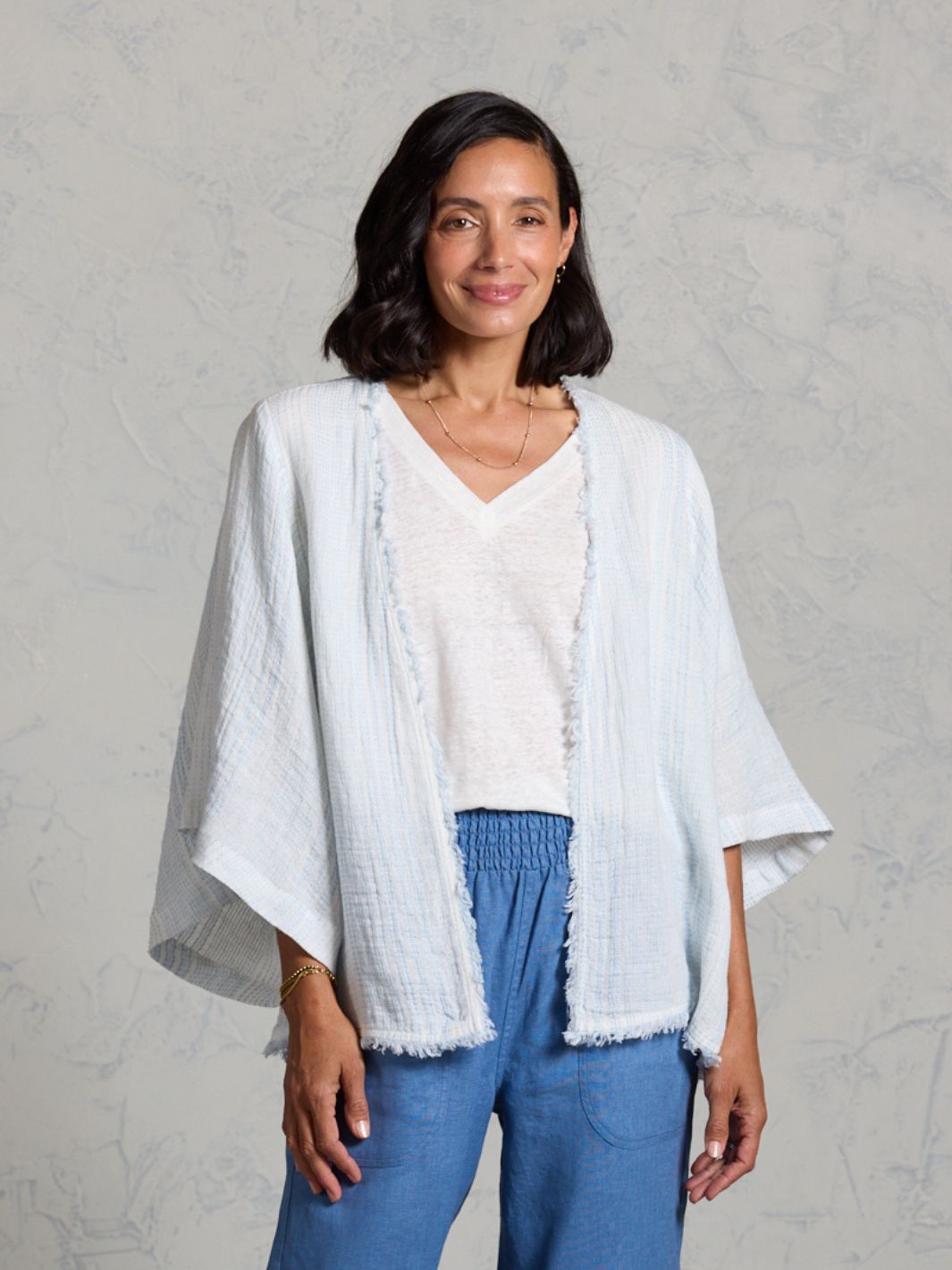 Woman wearing a light blue kimono-style garment over a white top and blue pants against a gray background