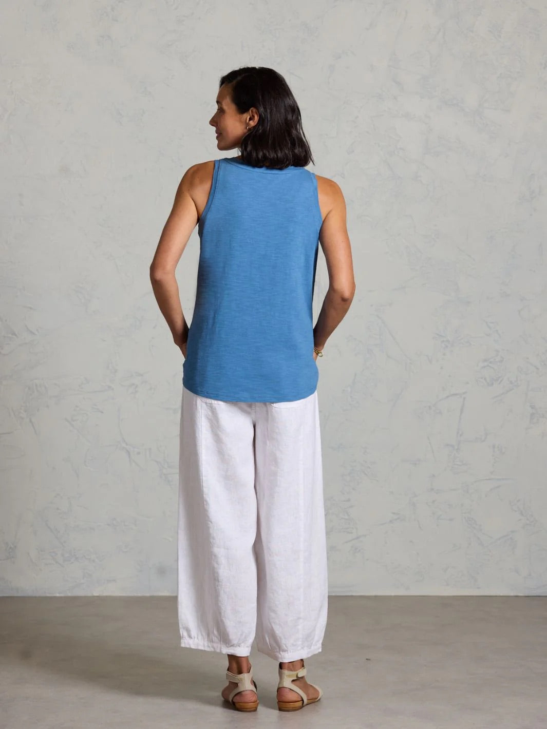 Woman wearing a blue sleeveless top and white pants against a gray background